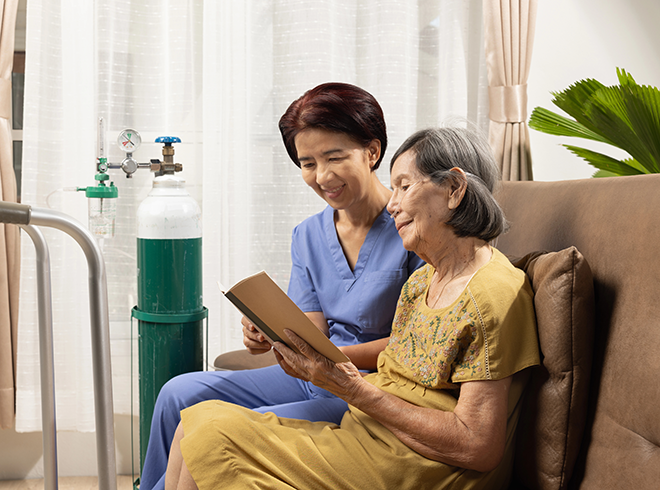 Home healthcare nurse reading with elderly woman on oxygen therapy, illustrating in-home chronic care support and compassionate patient engagement
