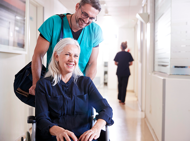 Healthcare provider assisting smiling older woman in wheelchair through clinic hallway, highlighting patient mobility support and transitional care services