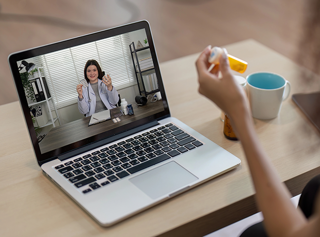 Patient holding prescription bottle during telehealth visit with doctor on laptop screen, illustrating virtual care delivery, medication reconciliation, and remote patient support