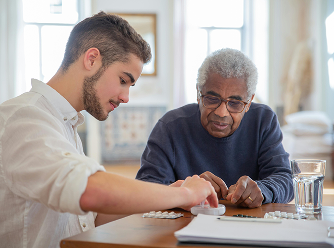 Young male caregiver assisting an elderly man with daily medication management at home, highlighting supportive chronic care in a senior-friendly setting